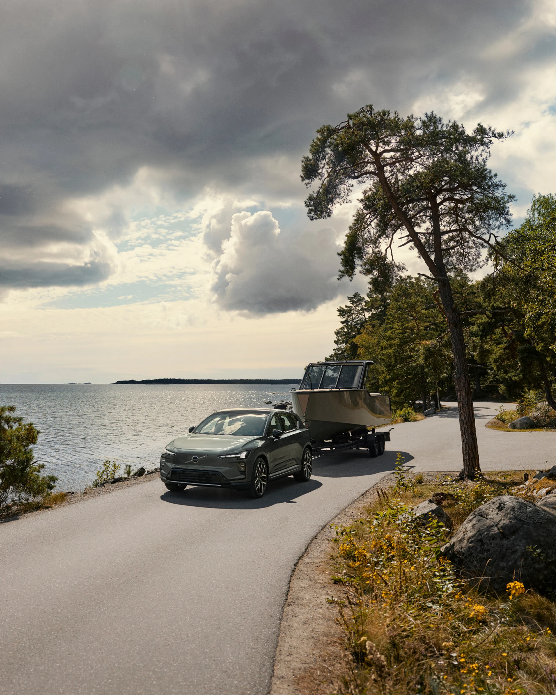 Volvo EX60 буксирует лодку во время движения по прибрежной дороге рядом с водой на фоне деревьев и облаков.