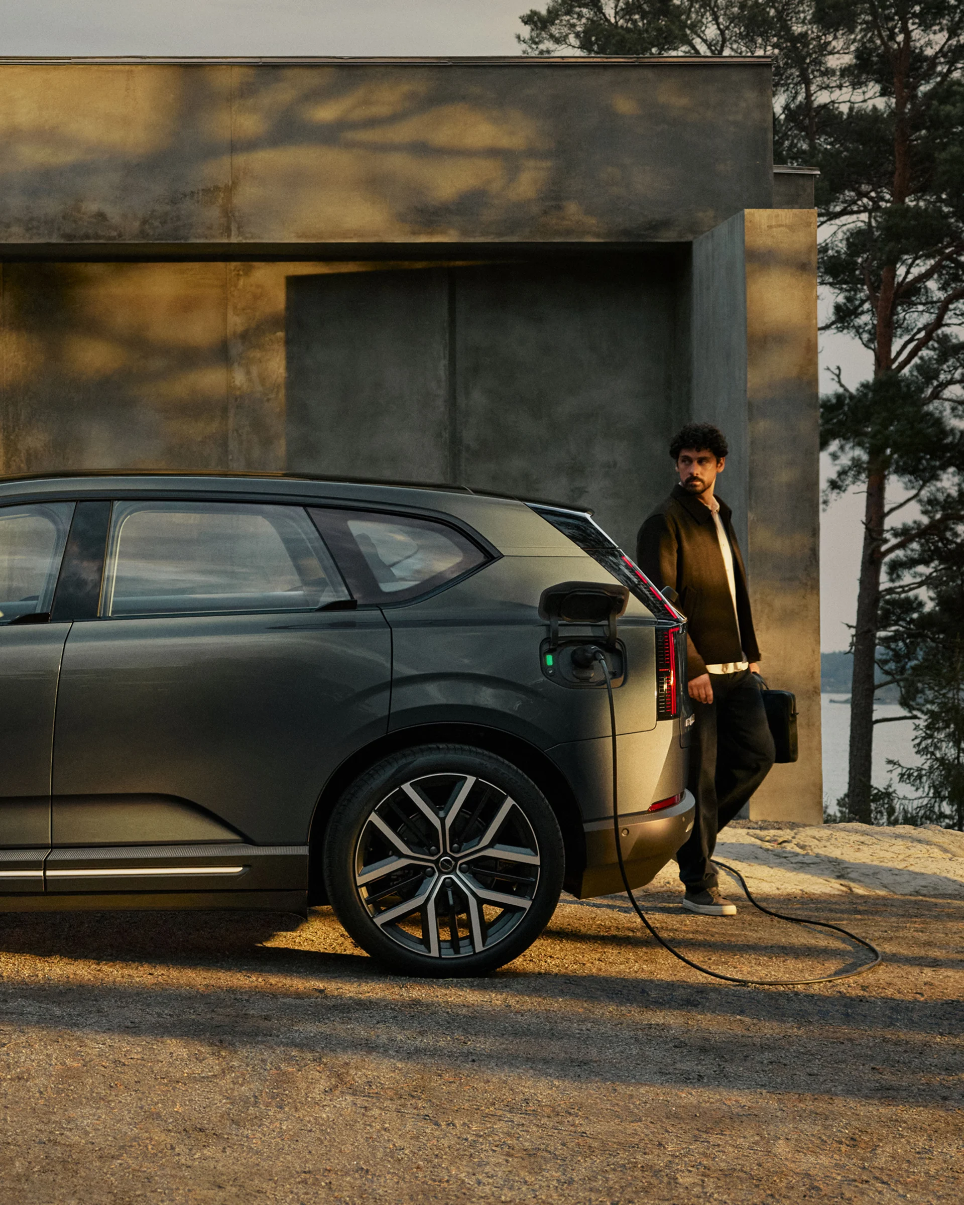 Человек стоит рядом с Volvo EX60, подключенным к домашнему зарядному устройству в сумерках.