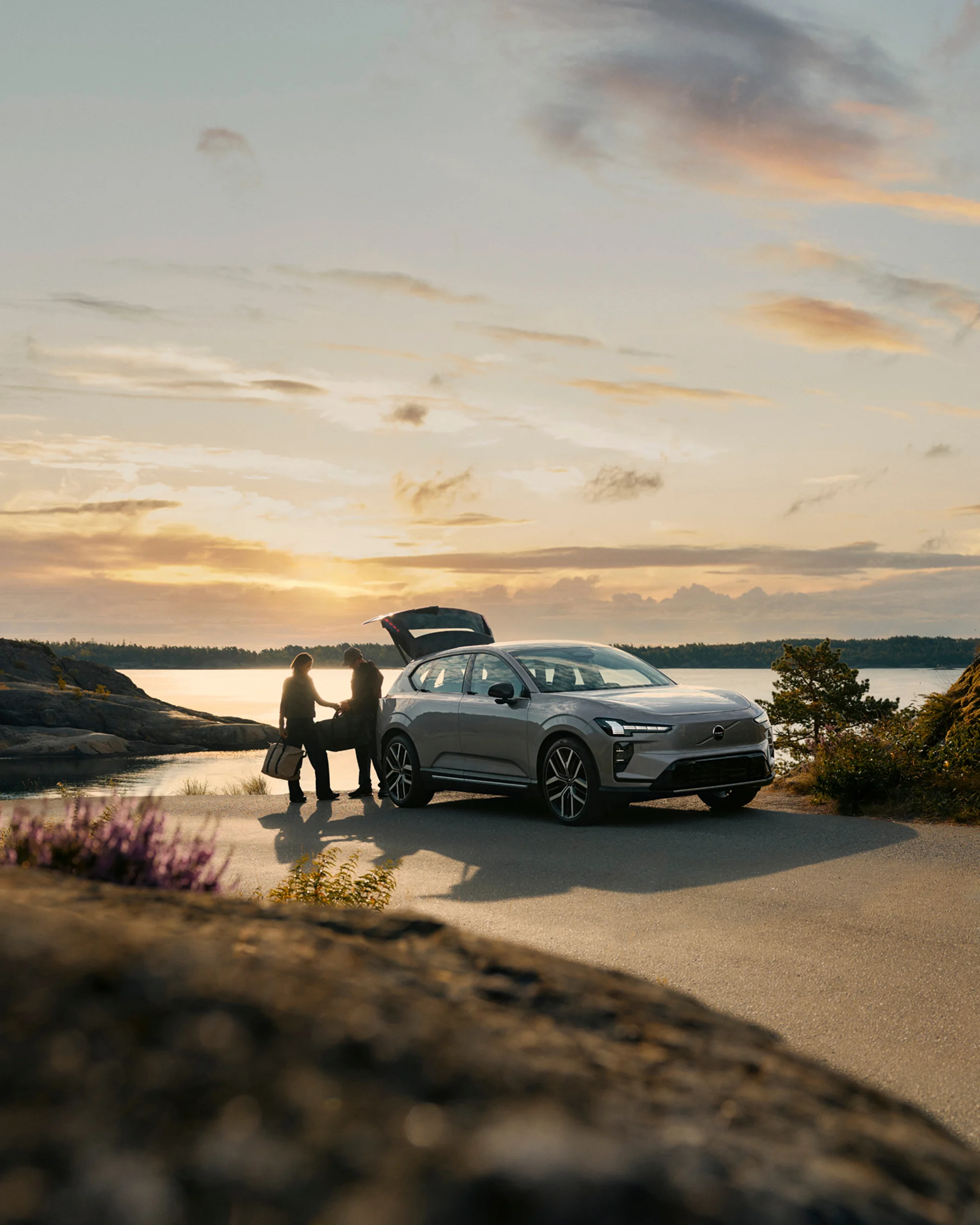 Volvo EX60 припаркован у берега на закате с двумя людьми, выгружающими снаряжение из открытого багажника.