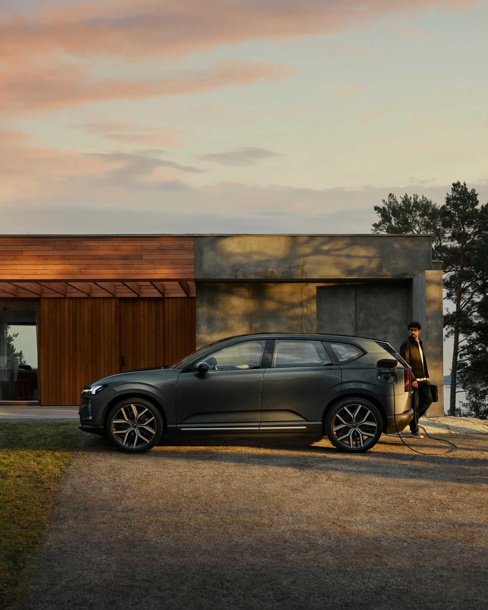 Volvo EX60 заряжается у припаркованного у современного дома, вид сбоку, позади идет человек.