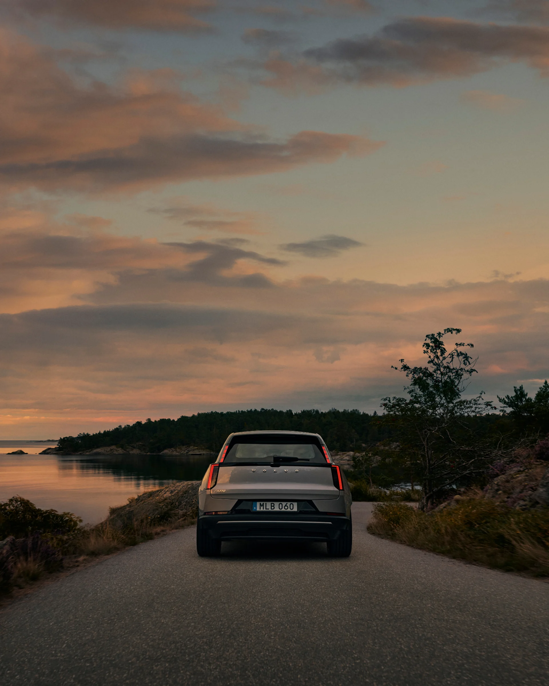 Вид сзади на Volvo EX60, едущий по прибрежной дороге в сумерках.