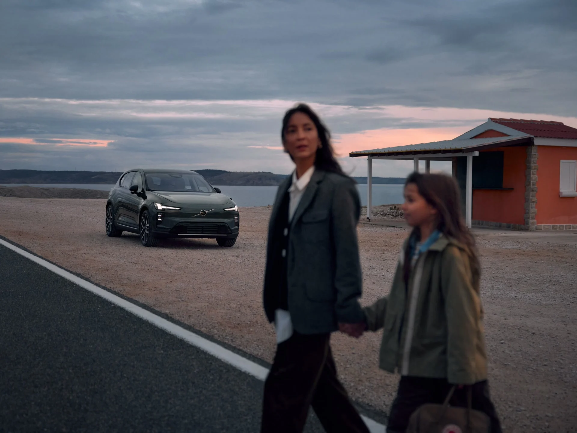 На заднем плане резко виден электромобиль Volvo, а на переднем плане — размытая женщина, держащая ребенка за руку.