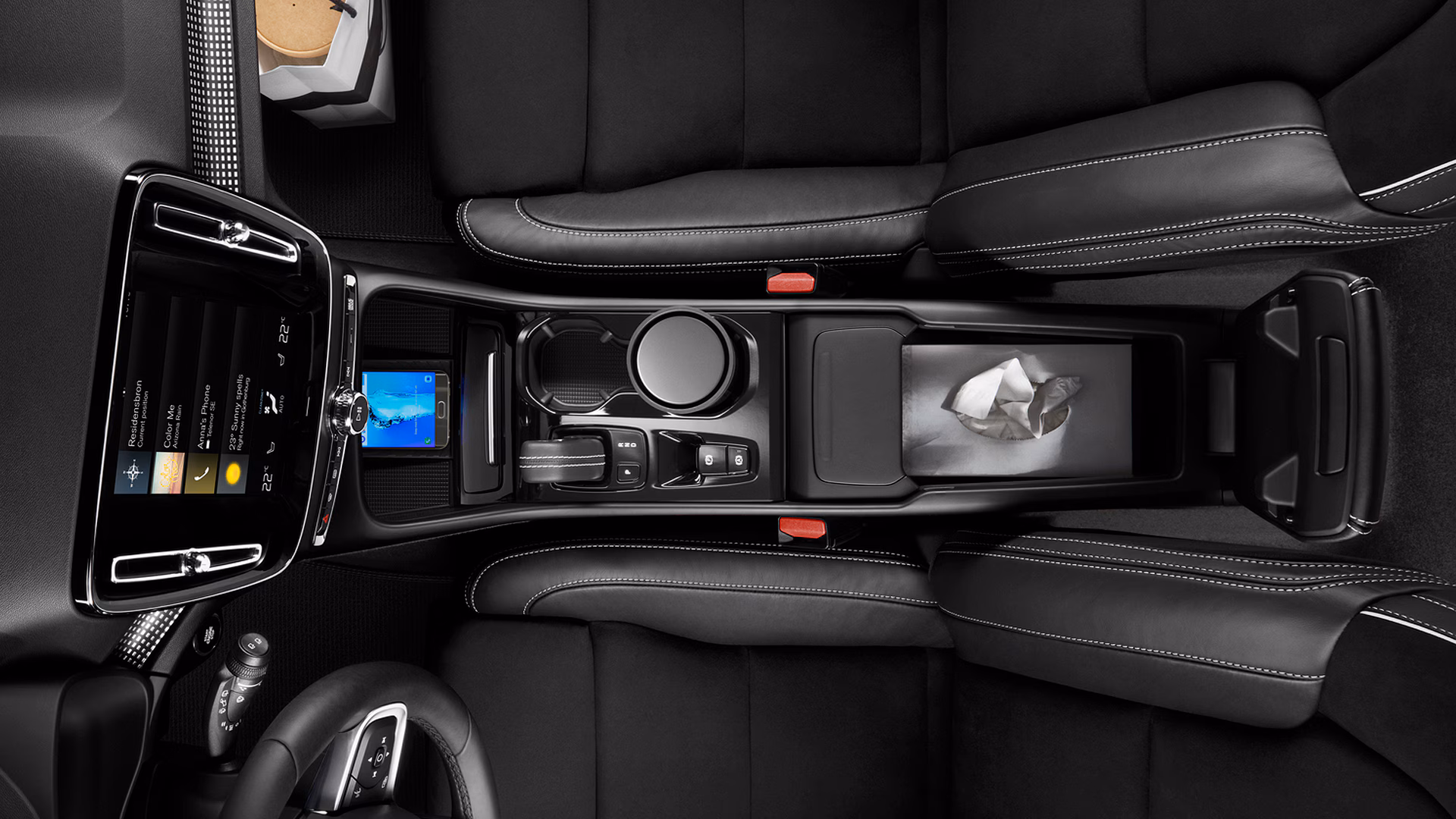 Вид сверху на центральную консоль салона автомобиля Volvo с информационно-развлекательным экраном, селектором передач, подстаканниками, слотом для смартфона и отсеком для хранения.