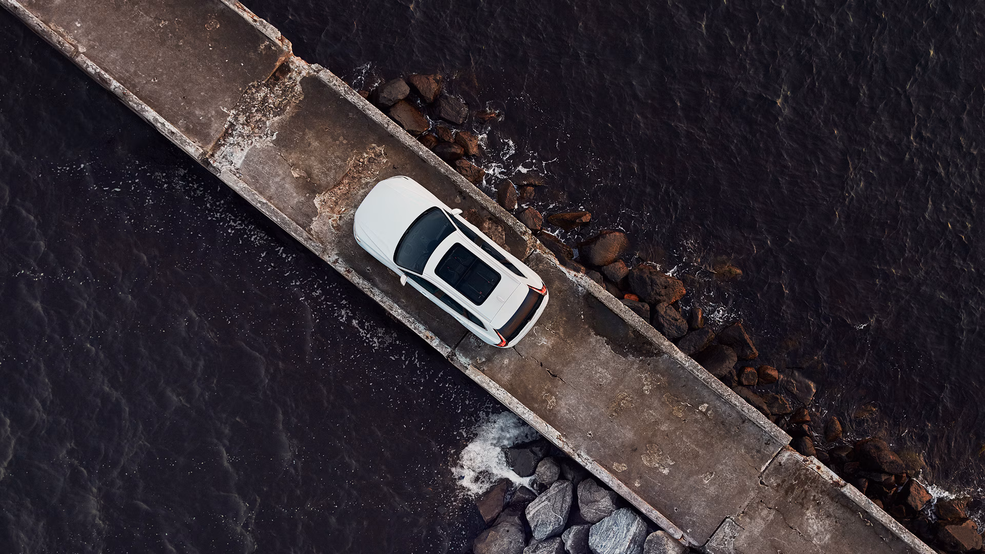 Вид сверху на белый автомобиль Volvo, припаркованный на узком бетонном пирсе рядом с темной водой и камнями.
