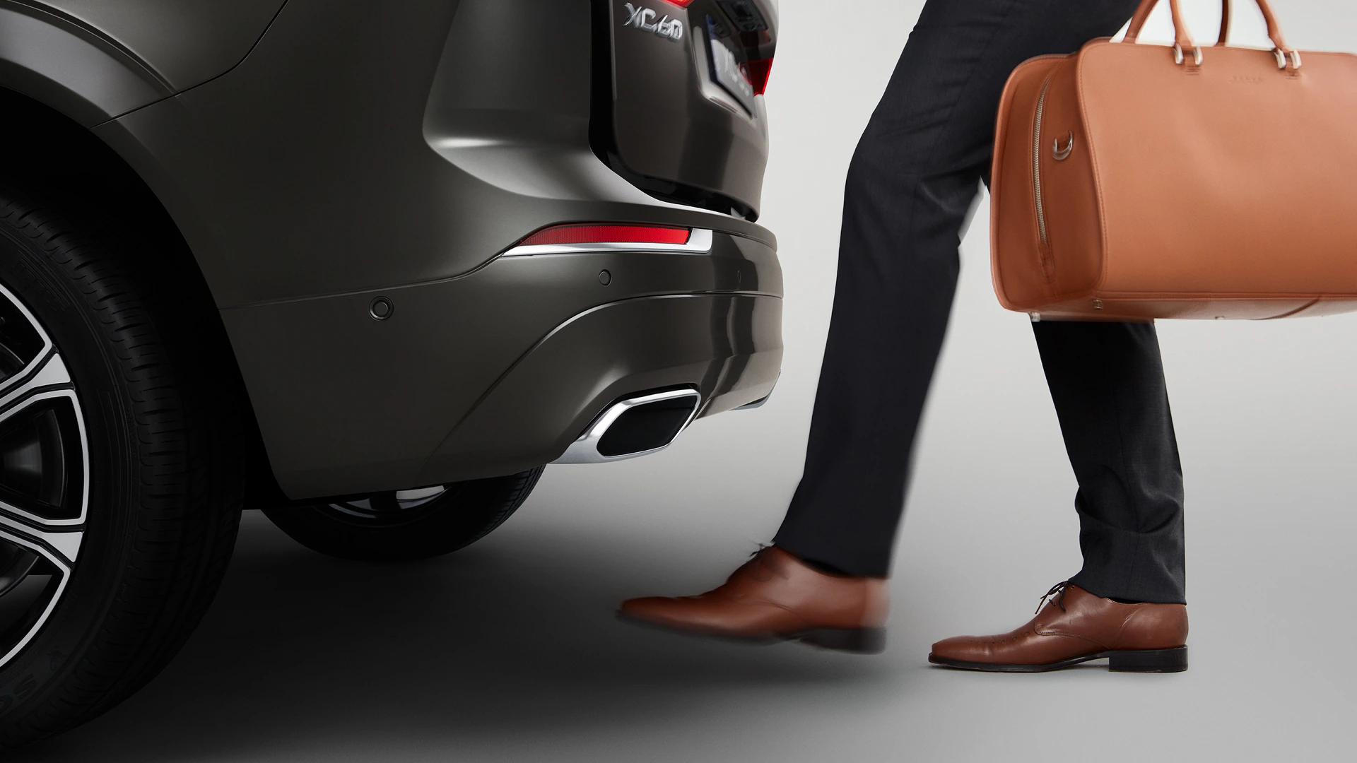 Человек с кожаной сумкой открывает заднюю часть Volvo XC40 с помощью датчика, активируемого ногой.