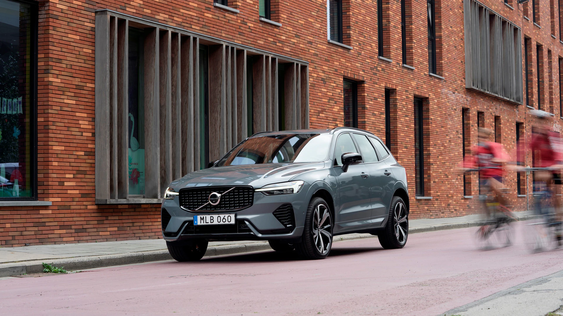 Volvo XC60 припаркован на городской улице рядом с кирпичным зданием, мимо которого едут два велосипедиста.