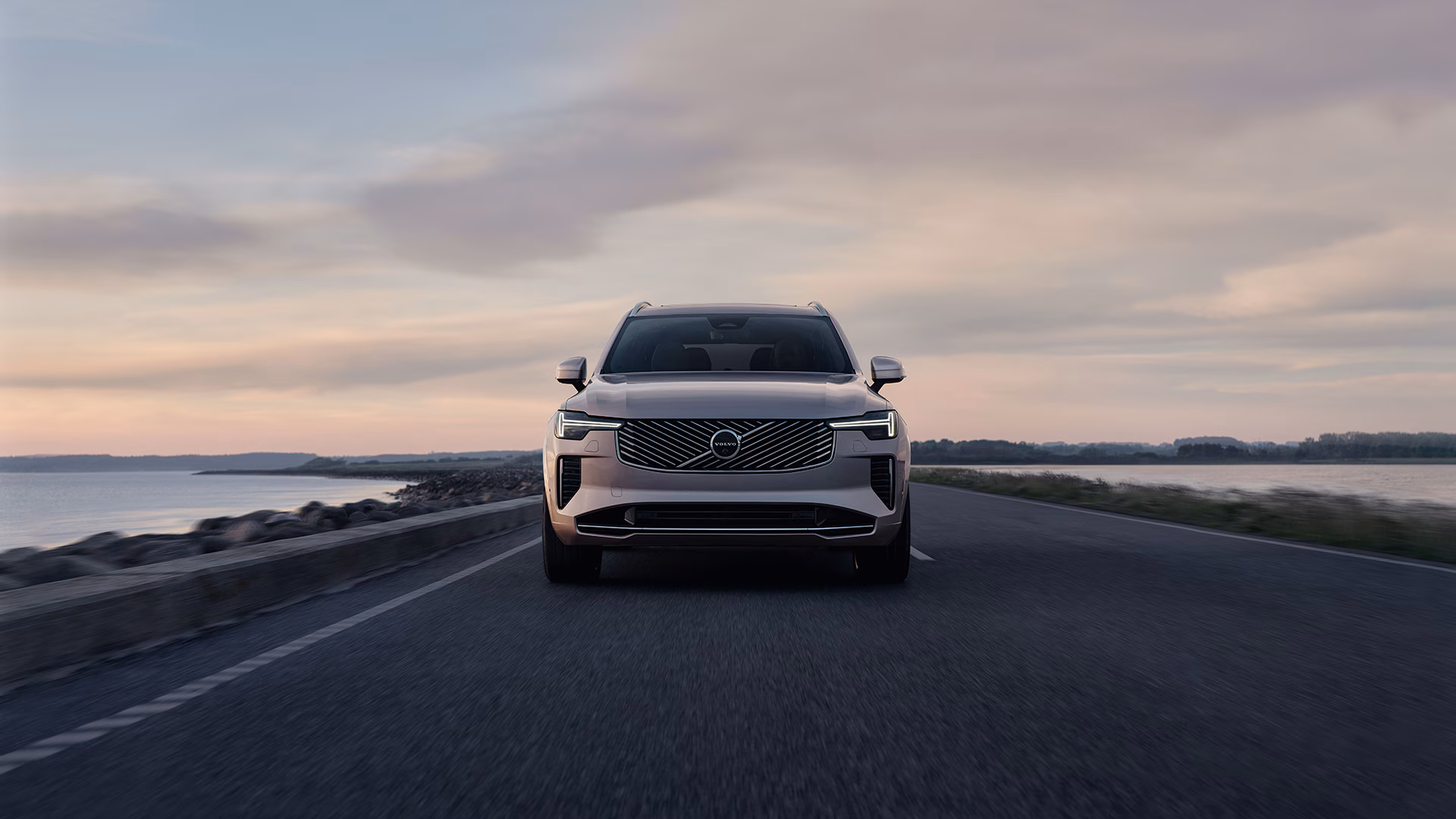 Вид спереди на внедорожник Volvo, едущий по прибрежной дороге на закате.