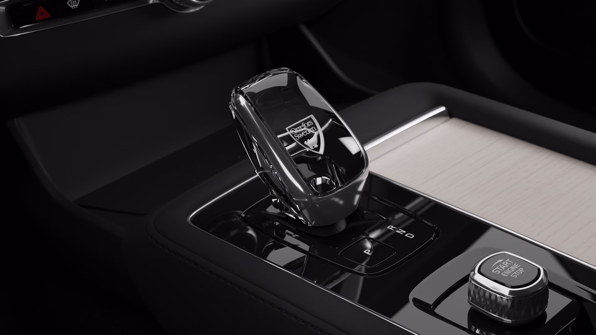 Крупный план хрустального переключателя передач автомобиля Volvo с гравировкой «Orrefors Sweden» рядом с кнопкой остановки двигателя на центральной консоли.